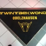 Dreieckige Logomatte mit gelbem Logo und Schriftzug in Bodenaussparung eingepasst
